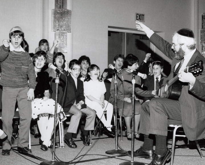 Black and white photo from 1980s showing a man playing a guitar and kids singing.