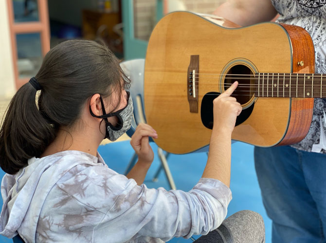 Student strumming guitar with one finger as music instructor holds it.