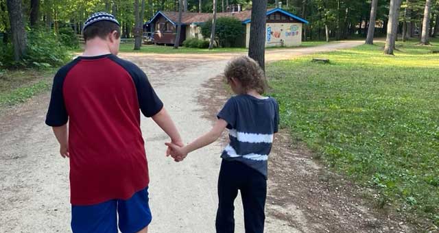 A boy wearing a kipah holding hands with a girl as they walk down a path at camp