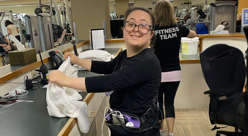 Adult team member standing behind a front desk and folding a towel. She's looking at the camera and smiling