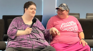 Lily and Sarah sitting in chairs and holding hands while Lily holds a microphone