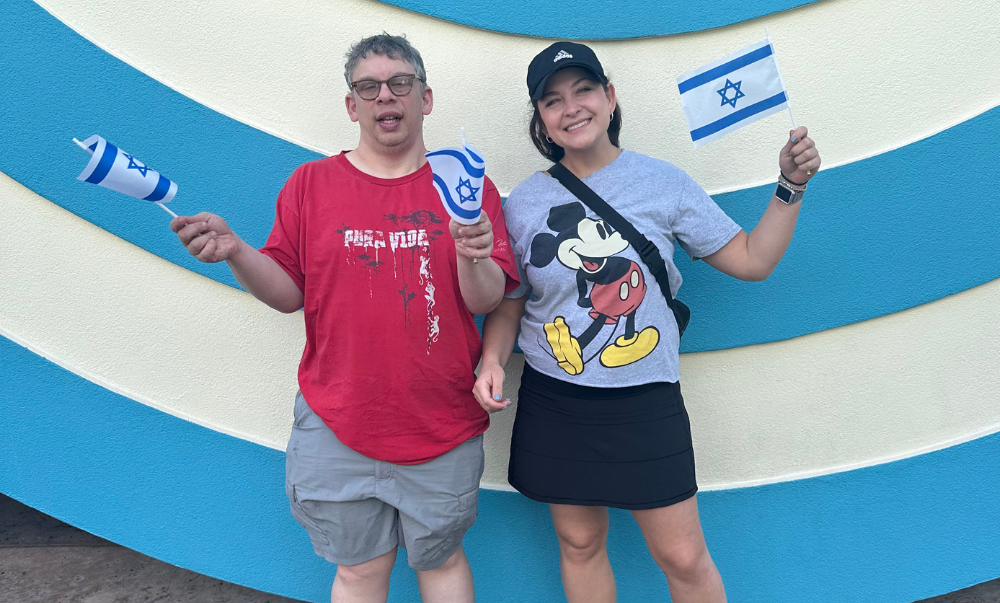 Two adults in front of a blue and white background holding Israeli flags