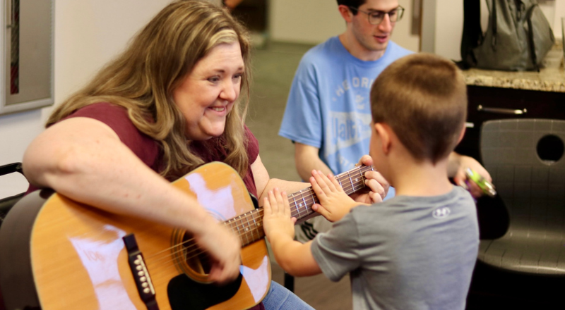 Music therapist strumming a guitar while a Keshet participant holds the guitar with her